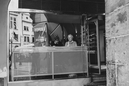 Černobílá pouliční fotografie zachycující tři ženy uvnitř kavárny přes prosklenou výlohu.
