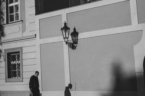 Černobílá street fotografie muže procházejícího kolem historické budovy s lucernou.
