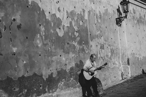 Černobílá street fotografie pouličního hudebníka hrajícího na kytaru u historické zdi v Praze.